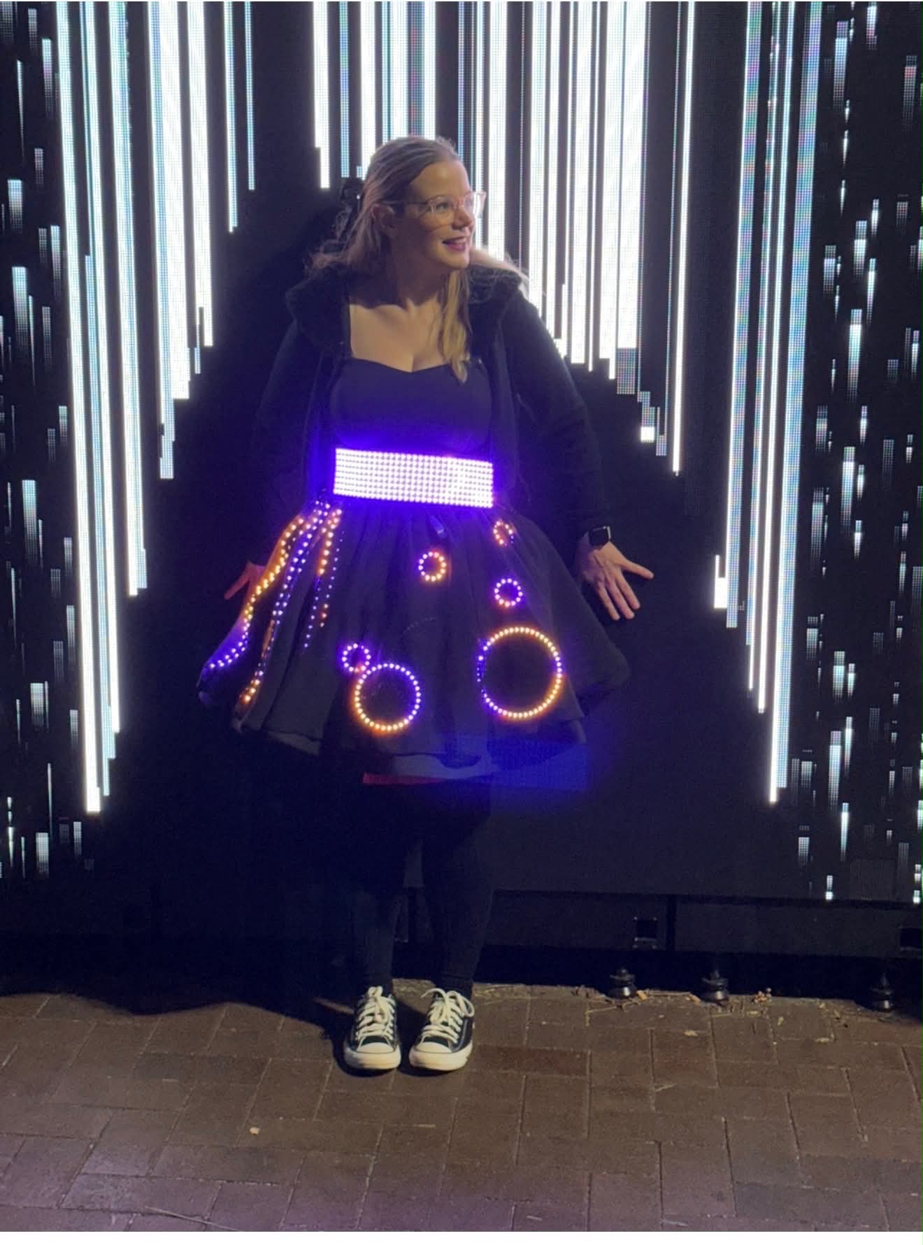 Kate Kirwin in her black made-to-measure dress, the skirt lit with rings and strips of violet and amber LEDs, in front of a wall of light