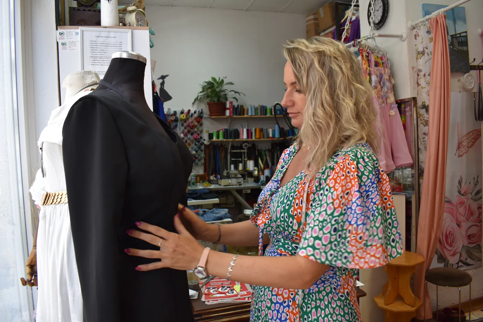Enisa fitting a black tailored blazer on a dress form in her studio, spools of thread on the shelves behind