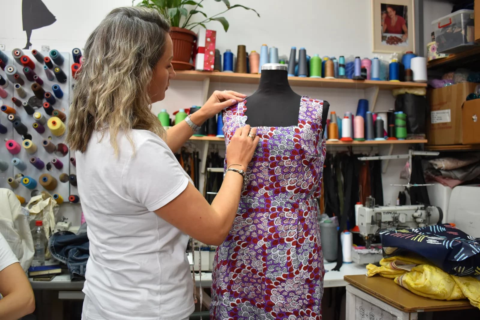 Enisa adjusting the strap of a purple printed MadeApt dress on a dress form, a sewing machine and thread cones beside her