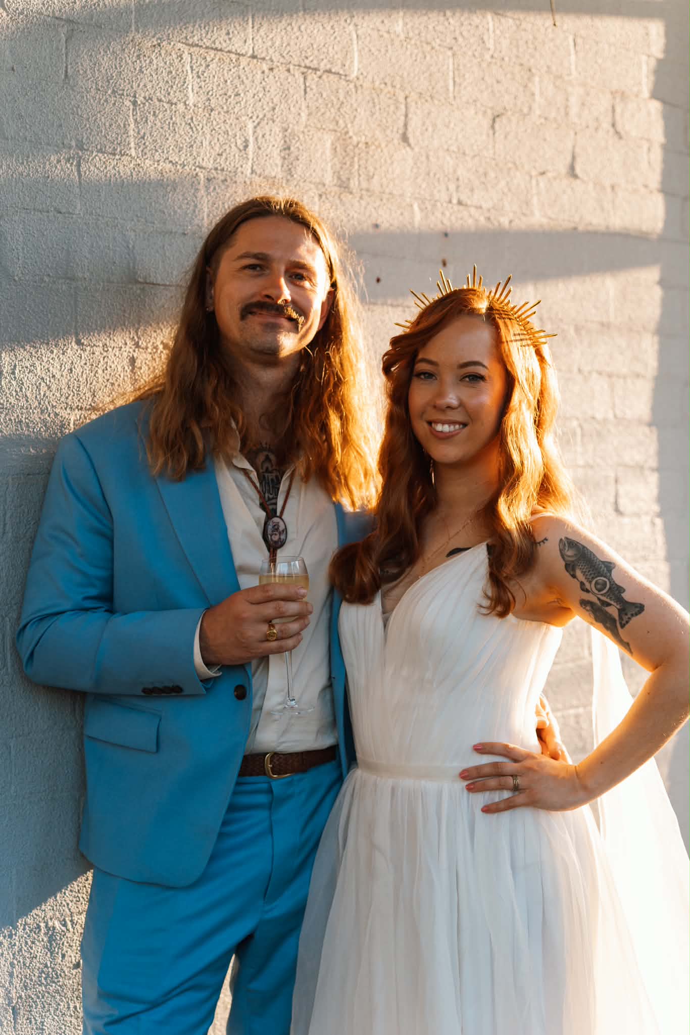 A bride in a white tulle V-neck gown and gold crown beside her partner in a blue suit at golden hour