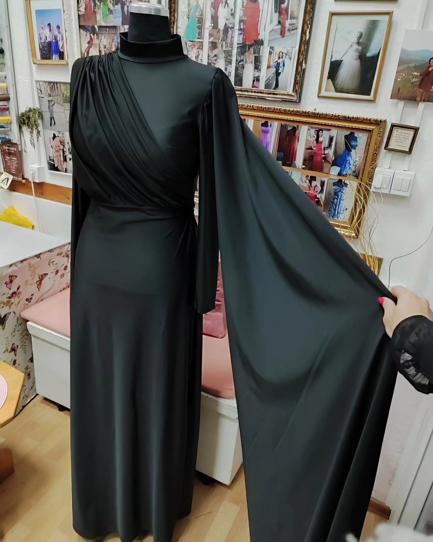 A custom black high-neck gown with a draped cape sleeve — an abaya-inspired modest silhouette in the tailor's studio