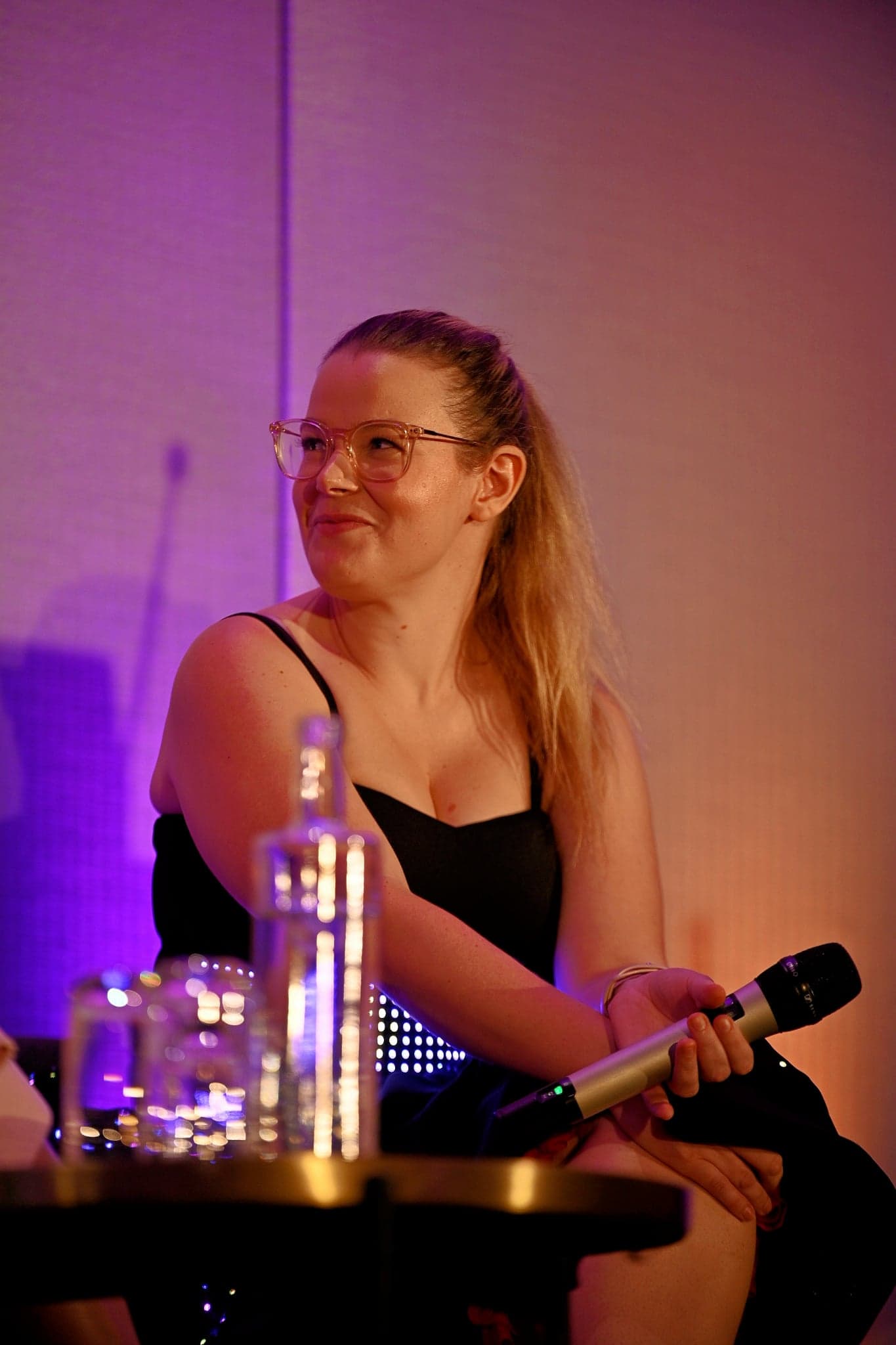 Kate Kirwin on a panel, holding a microphone, in the black sweetheart-neck dress with its lights off