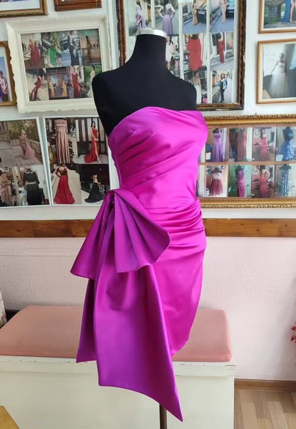 A fuchsia satin dress with a sculptural bow taking shape on a mannequin in a tailor's studio — a real garment in progress