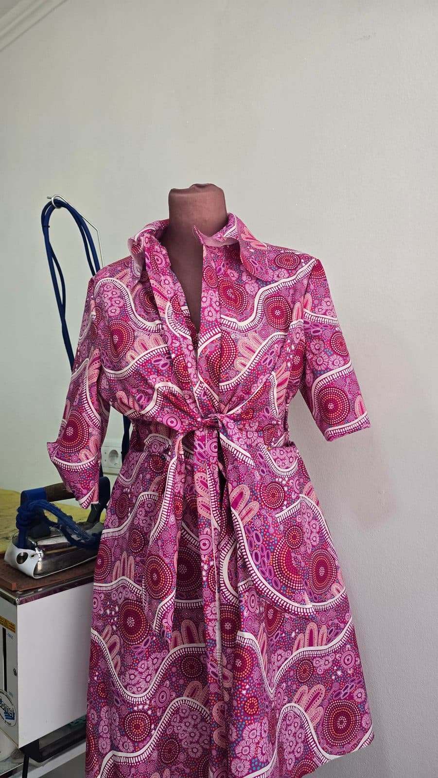 A magenta print shirt dress with a tie front on a dress form beside a steam iron