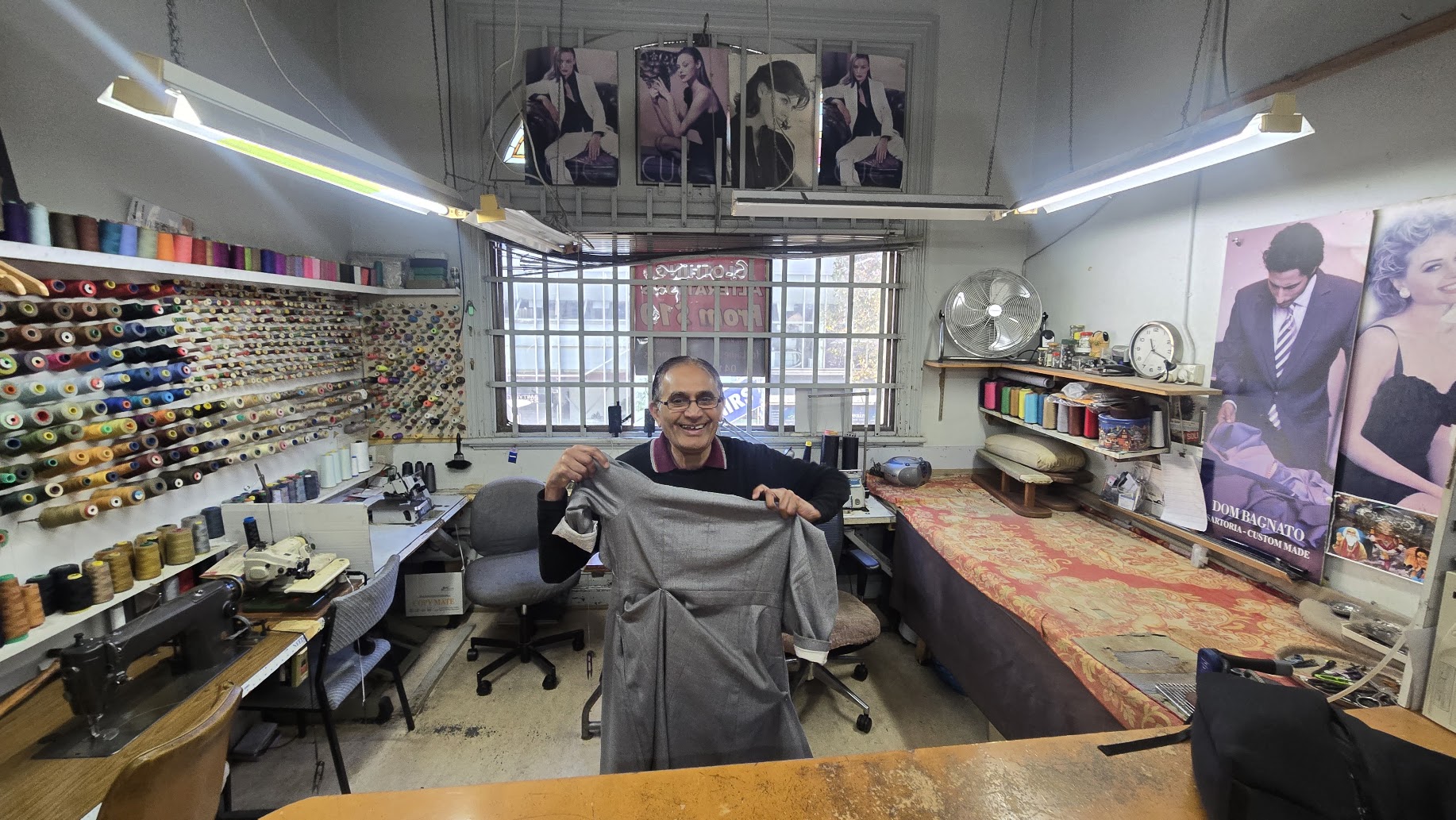A master tailor in his workroom holding up a grey garment in progress